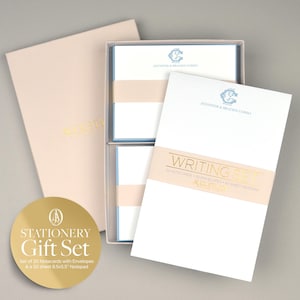 Pode incluir: Um conjunto de papelaria de presente numa caixa rosa claro com um logotipo dourado. O conjunto inclui 20 cartões com envelopes e um bloco de notas de 50 folhas. Os cartões são brancos com uma borda azul e um monograma dourado. O bloco de notas é branco com uma borda dourada e as palavras "Writing Set" em dourado.