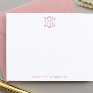 Puede incluir: Una tarjeta de papelería blanca con un monograma rosa y el nombre "ELIZABETH SANDERSON" impreso en rosa. También se ven un sobre rosa, clips dorados y un bolígrafo dorado.