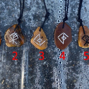 Könnte beinhalten: Fünf Bernstein-Anhänger mit verschiedenen Runen, die in sie geschnitzt sind. Jeder Anhänger ist an einer schwarzen Schnur befestigt und von 1 bis 5 nummeriert.