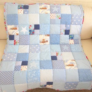 Może przedstawiać: Niebieska patchworkowa kołdra z różnymi wzorami, w tym gwiazdami, kropkami i misiami. Kołdra składa się z kwadratów w różnych odcieniach niebieskiego i bieli. Kołdra ma około 100 cm szerokości i 127 cm długości.