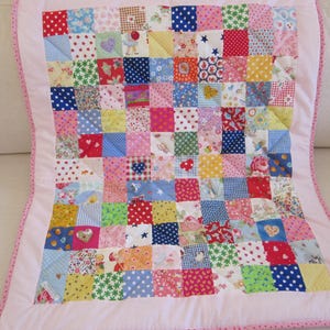 May include: A patchwork quilt with a pink border and backing. The quilt is made of numerous small, colorful squares of fabric, each with a different pattern, including polka dots, stars, and floral designs. The quilt is likely handmade.