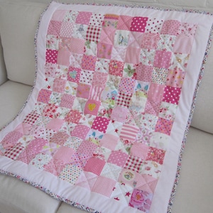 May include: A pink patchwork quilt with squares of various patterns, including polka dots, stripes, and floral prints. The quilt is bordered with white fabric and a floral trim. The quilt is made of various shades of pink.