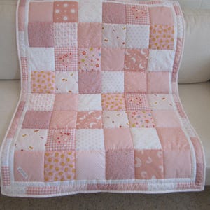 May include: A patchwork quilt featuring a grid of square fabric blocks in various shades of pink and white. The quilt displays diverse patterns including hearts, stars, moons, rainbows, and polka dots, finished with a solid white border trim.