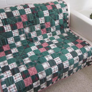 May include: A patchwork quilt in shades of green, white, and red, with a pink border. The quilt is arranged on a white sofa. The design is made up of squares.