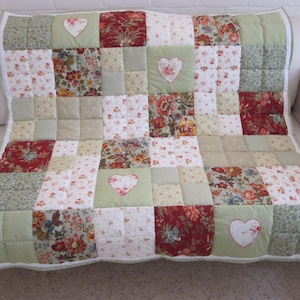 Könnte beinhalten: Eine Patchwork-Decke mit einem wiederkehrenden Muster aus salbeigrünen, dunkelroten und cremefarbenen Stoffquadraten. Das Design umfasst florale Drucke, Punkte und vier weiße herzförmige Applikationen, die auf die grünen Stoffteile genäht sind.