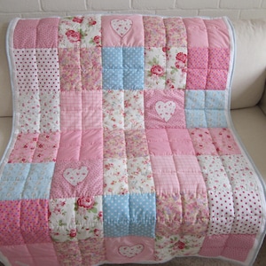 May include: A patchwork quilt in shades of pink, white, and light blue, featuring floral and polka dot patterns, and heart appliqués. The quilt is displayed on a cream-colored sofa, showcasing its decorative design and soft texture.