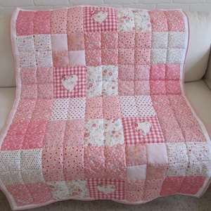 May include: A patchwork quilt in shades of pink and white, featuring various patterns like polka dots, floral prints, and gingham. The quilt has a heart-shaped appliqué and is displayed on a white sofa. The quilt is approximately 40 inches wide and 50 inches long.