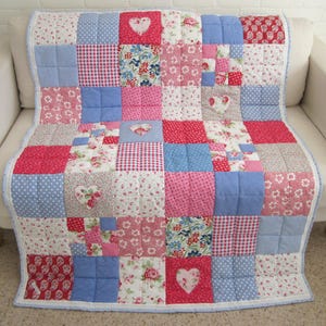 May include: A patchwork quilt in shades of red, pink, blue, and white, featuring floral, polka dot, and gingham patterns. Heart-shaped appliqués are sewn onto the quilt. The quilt is displayed on a white sofa, with a light blue border.
