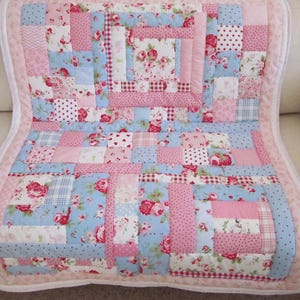 May include: A patchwork quilt featuring a variety of fabric squares in shades of pink and light blue. The design incorporates floral patterns, polka dots, gingham checks, and stripes, all bordered by a solid light pink fabric trim edge.