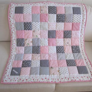 May include: A pink and grey patchwork baby quilt with a floral border. The quilt is made of squares of different fabrics, including pink, grey, white, and patterned fabrics.