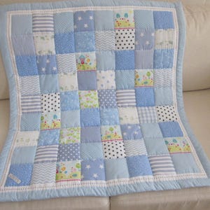May include: A patchwork quilt featuring a variety of light blue, white, and patterned fabric squares. The design includes prints of stars, stripes, chevrons, polka dots, and small landscape scenes with houses, all bordered by a solid light blue trim.