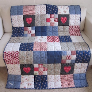 May include: A patchwork quilt with a variety of fabric patterns in red, white, and blue, including floral, polka dot, and striped designs. The quilt features three red heart appliqués on a black square. The quilt is draped over a white sofa.
