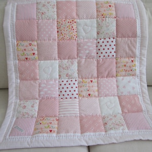 May include: A patchwork quilt featuring a grid of square fabric patches in various shades of pink and white. The design includes patterns of stars, hearts, stripes, and floral motifs, with quilted heart shapes stitched into select white squares.
