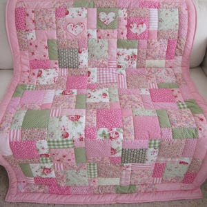 May include: A patchwork quilt in shades of pink and green, featuring floral and polka dot patterns. The quilt has a pink border and includes heart-shaped appliqués. It is draped over a white sofa.