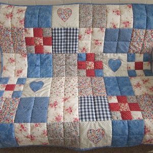 Könnte beinhalten: Ein Patchwork-Quilt mit blauem Rand, mit Quadraten aus floralen, Vichy- und einfarbigen blauen Stoffen. Rote und weiße Quadrate und Herzapplikationen fügen Details hinzu. Der Quilt wird auf einem weißen Sofa präsentiert.