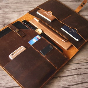 May include: A brown leather portfolio with multiple pockets and compartments. The portfolio is open and contains a tablet, a phone, a notebook, a pen, a watch, credit cards, and a passport.