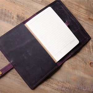 May include: A purple leather notebook cover with a blank lined notebook inside. The cover has a strap with gold metal snaps and a pocket on the back.