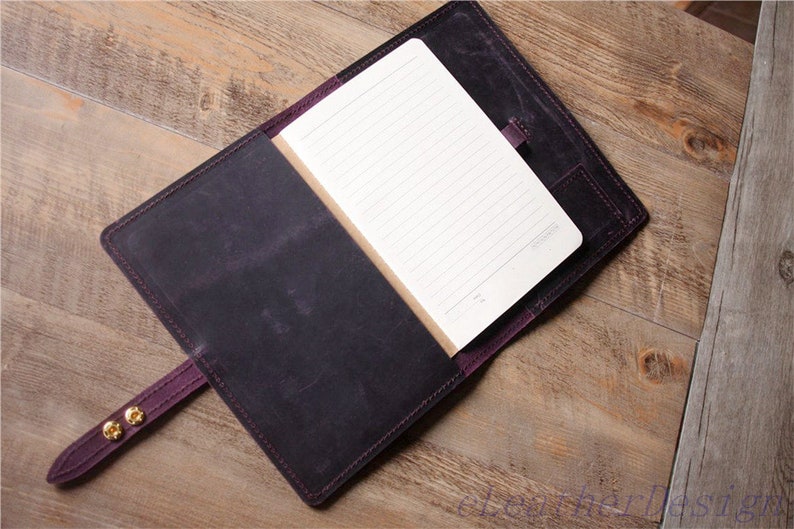 May include: A purple leather notebook cover with a blank lined notebook inside. The cover has a strap with gold metal snaps and a pocket on the back.