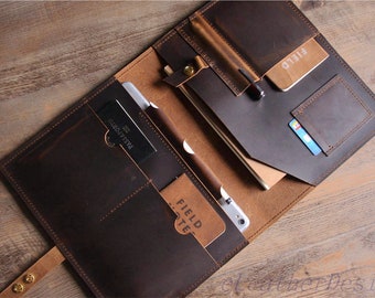 Pode incluir: Um portfólio de couro marrom com vários bolsos e compartimentos. O portfólio está aberto para revelar um caderno, uma caneta, um cartão de crédito e um passaporte. O texto "FIELD NOTE" é visível no interior do portfólio.