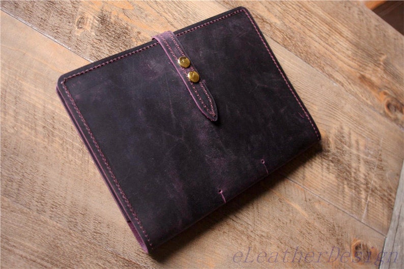 May include: A dark purple leather notebook with a strap and two gold metal clasps. The notebook has a distressed look and is stitched with purple thread.