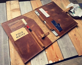 May include: A brown leather portfolio with multiple pockets and a snap closure. The portfolio contains a notebook, a pen, cards, and a passport.