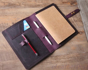 Peut inclure: Une couverture de carnet en cuir violet foncé avec une fente pour carte contenant une carte bleue, deux passants pour stylo avec un stylo rouge et argenté et un stylo blanc, et un carnet en papier beige. Fermeture à pression et texte eLeatherDesign.