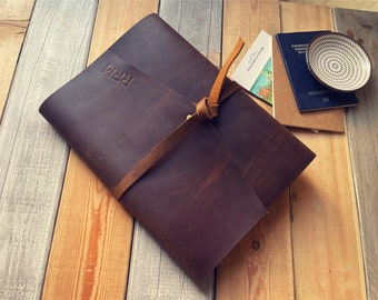 Peut inclure: Un carnet en cuir marron foncé avec une fermeture à lanière en cuir. La couverture avant présente les lettres RRM en relief. Le carnet est rectangulaire avec une finition mate lisse et un grain naturel visible, fixé par un nœud simple.