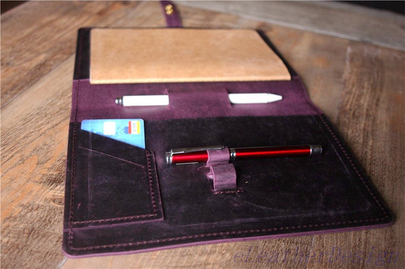 May include: A purple leather notebook cover with a pen holder and a card slot. The cover is open and there are two pens and a credit card inside.