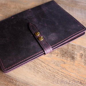 May include: A dark purple leather notebook with a strap and two gold metal clasps. The notebook has a stitched edge and is closed.