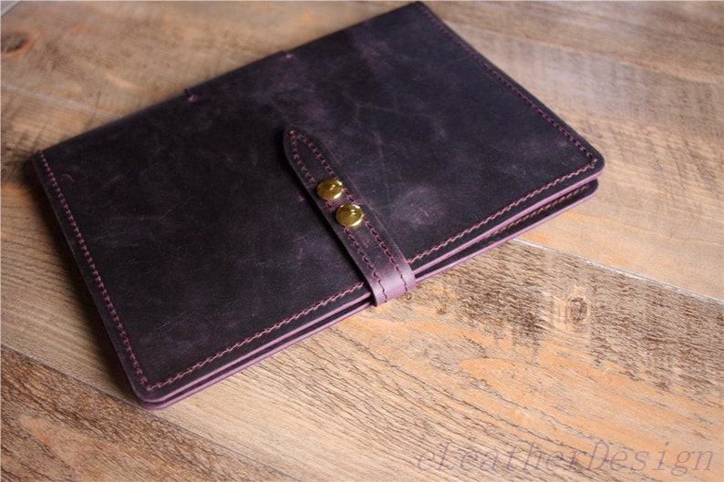 May include: A dark purple leather notebook with a strap and two gold metal clasps. The notebook has a stitched edge and is closed.