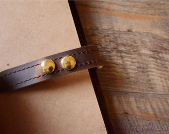 Peut inclure: Une sangle de fermeture en cuir marron foncé dotée de deux rivets métalliques dorés polis et de coutures apparentes, fixée sur une couverture de carnet en papier beige. La sangle maintient le carnet avec une bande de cuir lisse et solide.