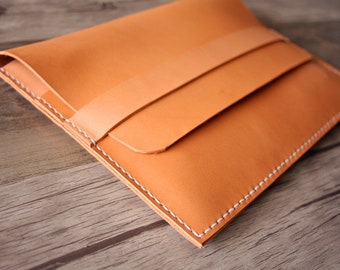 May include: A light brown leather laptop sleeve with a flap closure and white stitching. The sleeve is made of high-quality leather and has a minimalist design.