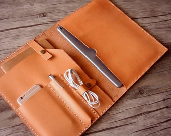 May include: A tan leather portfolio with multiple pockets and compartments. The portfolio is open and shows a tablet, phone, pen, and headphones.