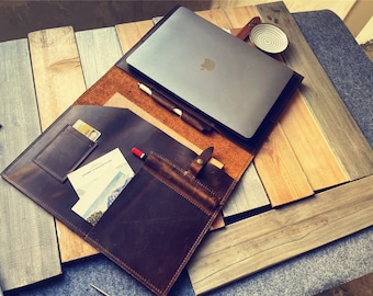 Pode incluir: Uma capa para laptop de couro marrom com vários bolsos e fecho de aba. A capa está aberta e um laptop, uma caneta e um cartão são visíveis no interior.