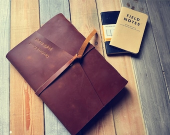 Pode incluir: Um diário de couro marrom com uma alça de couro bege e as palavras "Field Notes" gravadas na capa. O diário está sobre uma superfície de madeira.