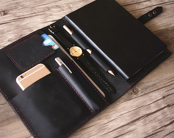 May include: Black leather portfolio with red stitching, open to reveal pockets for a phone, credit cards, pens, and a watch. The portfolio also has a notebook insert.