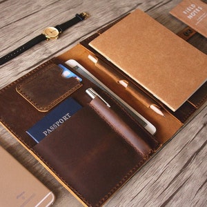May include: A brown leather travel journal with a passport holder, pen holder, and a notebook. The journal has a flap that closes with a snap.