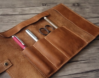 Pode incluir: Um rolo de couro marrom para canetas com cinco ranhuras para canetas e um pequeno bolso para um lápis ou outro item pequeno. O rolo está aberto e contém três canetas e um par de tesouras.