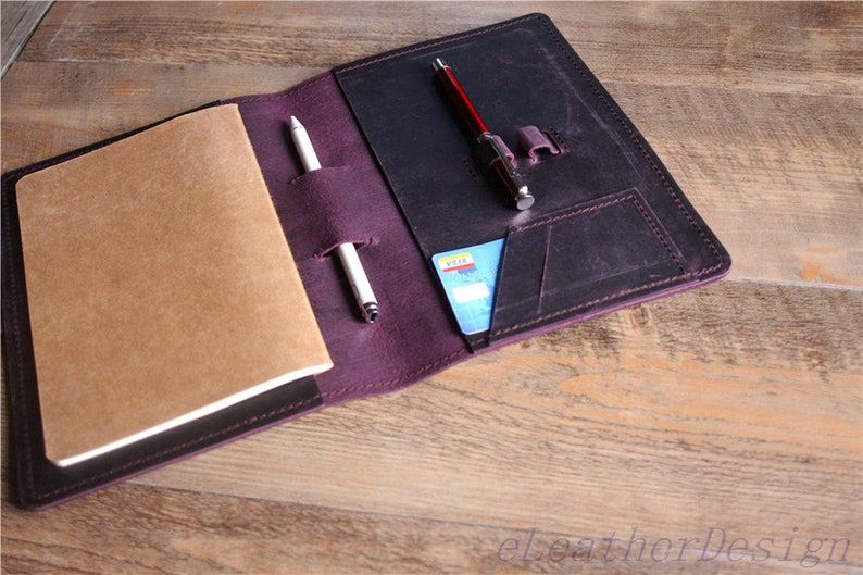 May include: A purple leather notebook cover with a brown notebook, a pen, and a credit card inside. The cover has a pocket for a pen and a slot for a credit card.