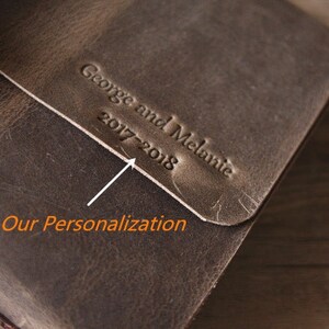 May include: A brown leather journal with the text "George and Melanie 2017-2018" embossed on the cover. The text is highlighted with a white arrow and the words "Our Personalization" are written below the arrow.