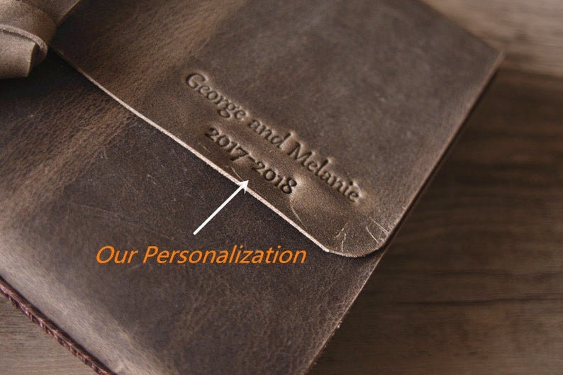May include: A brown leather journal with the text "George and Melanie 2017-2018" embossed on the cover. The text is highlighted with a white arrow and the words "Our Personalization" are written below the arrow.