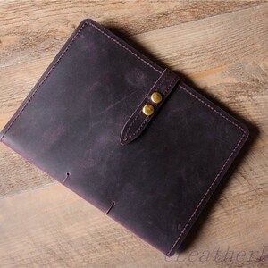 May include: A dark purple leather journal with a strap closure and two gold metal buttons. The journal has a distressed look and is stitched with purple thread.