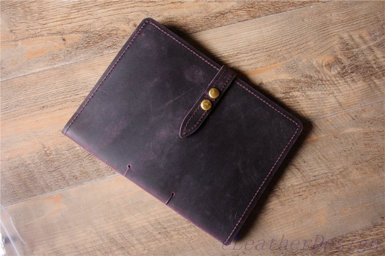 May include: A dark purple leather journal with a strap closure and two gold metal buttons. The journal has a distressed look and is stitched with purple thread.