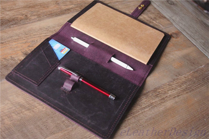 May include: A dark purple leather notebook cover with a brown paper notebook, a white pen, a red pen, and a credit card in the pocket.