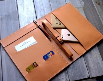 May include: A tan leather folio organizer featuring white stitching, a pen loop with a black and silver pen, two card slots containing credit cards, a business card, a leather wrist strap, and a pocket holding a Field Notes notebook and a smartphone.