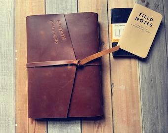 May include: A brown leather-bound journal with a strap closure and the text "NESTE Caroline & Pascal 349 N 842 W" embossed on the front cover. The journal is sitting on a wooden surface next to a black Moleskine notebook and a Field Notes notebook.