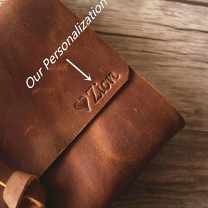 May include: A brown leather journal with a strap closure. The journal is personalized with the name "Zion" in a heart shape.