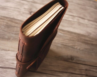 May include: A dark brown leather journal with a wrap-around strap closure. The notebook features visible stitching along the spine and contains multiple stacks of cream-colored paper pages visible from the top edge view.