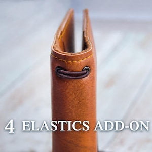 Könnte beinhalten: Ein Notizbuchumschlag aus hellbraunem Leder mit einem dunkelbraunen Gummiband, das durch eine Metallöse am Buchrücken geführt ist, sowie gelben Nähten an den Kanten. Der Text 4 ELASTICS ADD-ON ist unten im Bild in Weiß aufgedruckt.