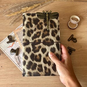 May include: A journal with a leopard print cover and a dragonfly clasp. The journal is open, displaying a map and vintage paper. Decorative tape and metal binder clips are on a wooden surface.